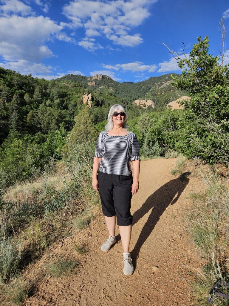 Linnea Mellinger smiles while standing on a hiking trail surrounded by pine-covered hills and red rock formations beneath a bright blue sky with scattered clouds.