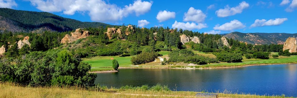 **Alt text:**

Panoramic view of Perry Park featuring a calm lake in the foreground, a lush green golf course winding through the landscape, and rugged red and tan rock formations surrounded by dense pine-covered hills beneath a bright blue sky with scattered white clouds.
